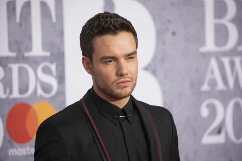 Liam Payne in a black suit jacket and black dress shirt poses against a grey backdrop 