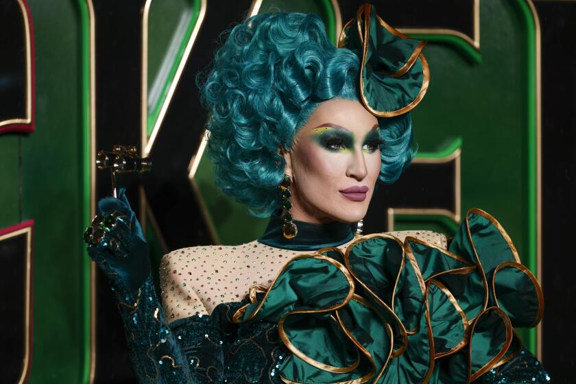 The Vivienne in an emerald green wig and a matching gown standing in front of a emerald green backdrop