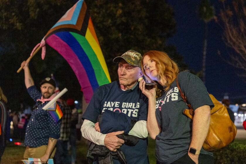 Riverside, CA - December 19: Transgender athlete supporter Kyle Harp, left, of Riverside holds the progress pride flag as "Save Girls Sports" supporters Lori Lopez and her dad Pete Pickering, both of Riverside, listen to the debate as they join the overflow crowd converging outside the Riverside Unified School District meeting Thursday night to debate the rights of transgender athletes to compete in high school sports Thursday, Dec. 19, 2024. (Allen J. Schaben / Los Angeles Times)