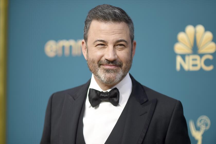 Jimmy Kimmel with greying facial hair smiles and poses in a black tuxedo against a turquoise backdrop with gold text