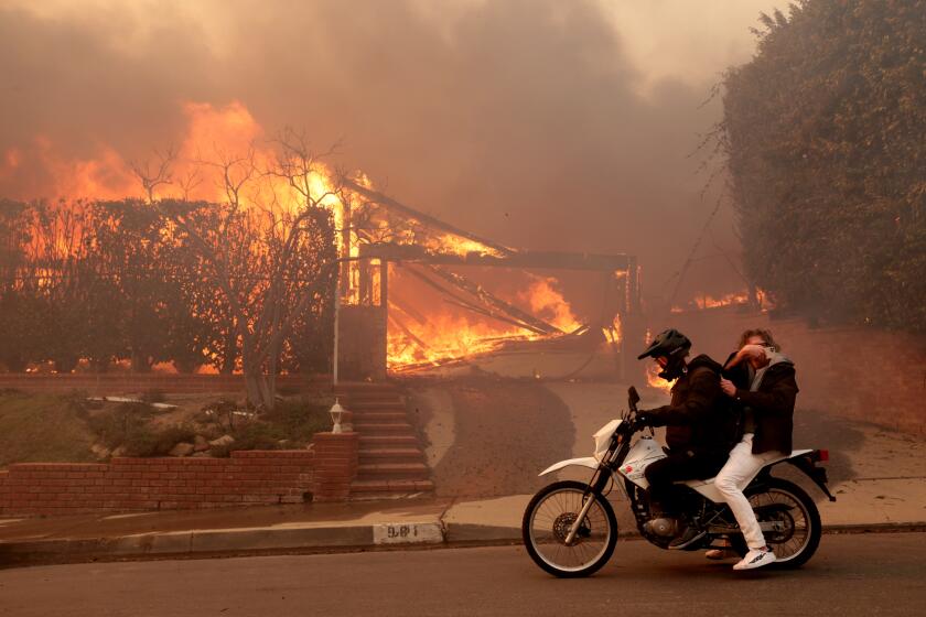 \wo people ride past a burning house off Enchanted Way in the Marquez Knolls neighborhood of Pacific Palisades.