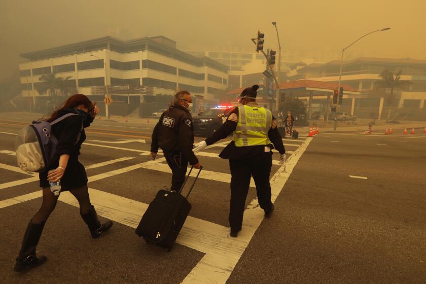 PACIFIC PALISADES, CA - JANUARY 7, 2025 - - An evacuee is helped across Pacific Coast Highway as smoke from the Palisades fire envelopes them at Sunset Blvd. In Pacific Palisades on January 7, 2025. (Genaro Molina/Los Angeles Times)