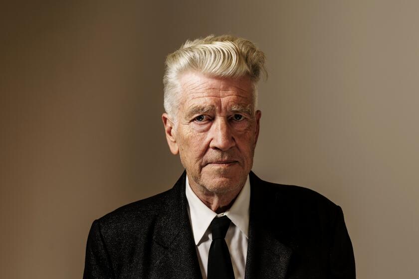 David Lynch in a suit with his hands to his sides posing against a brown background