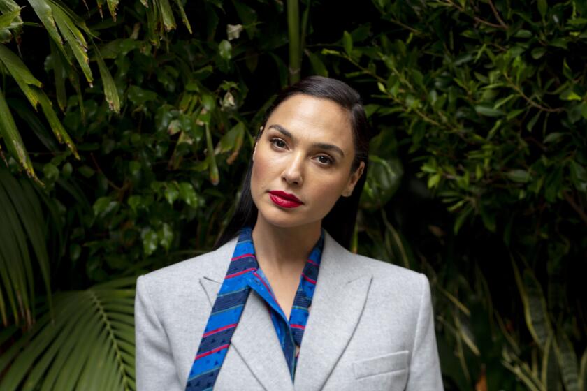  Gal Gadot poses in a gray blazer, blue blouse and striped pants in front of greenery